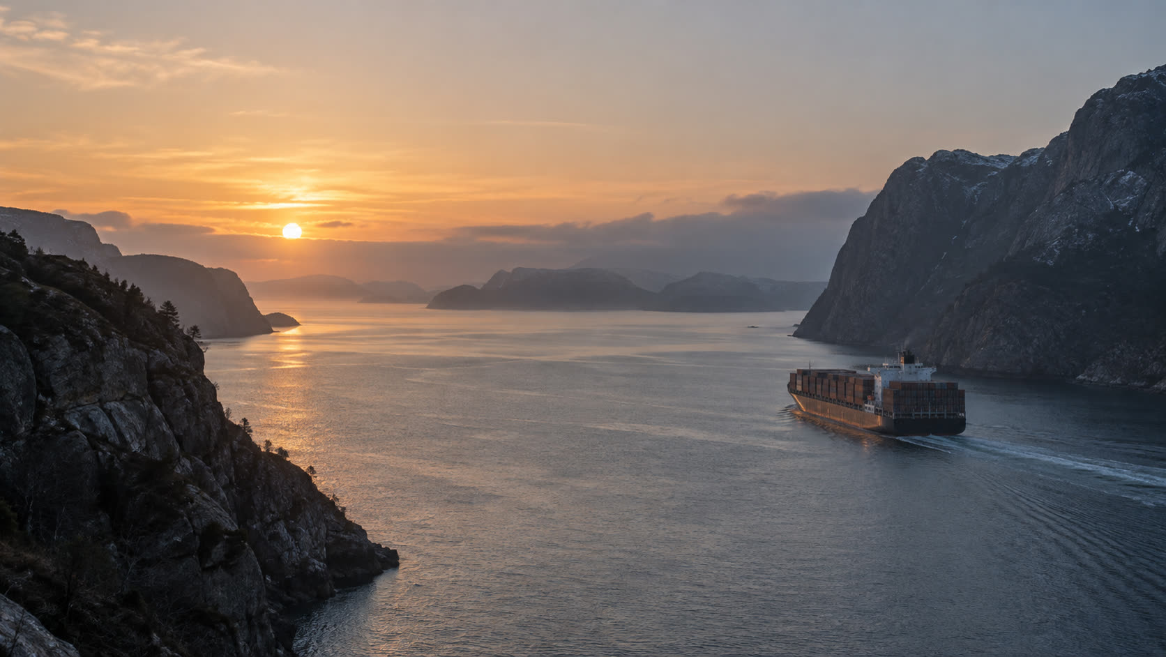 A container ship moving through a narrow sea strait between dark rocky headlands at sunrise